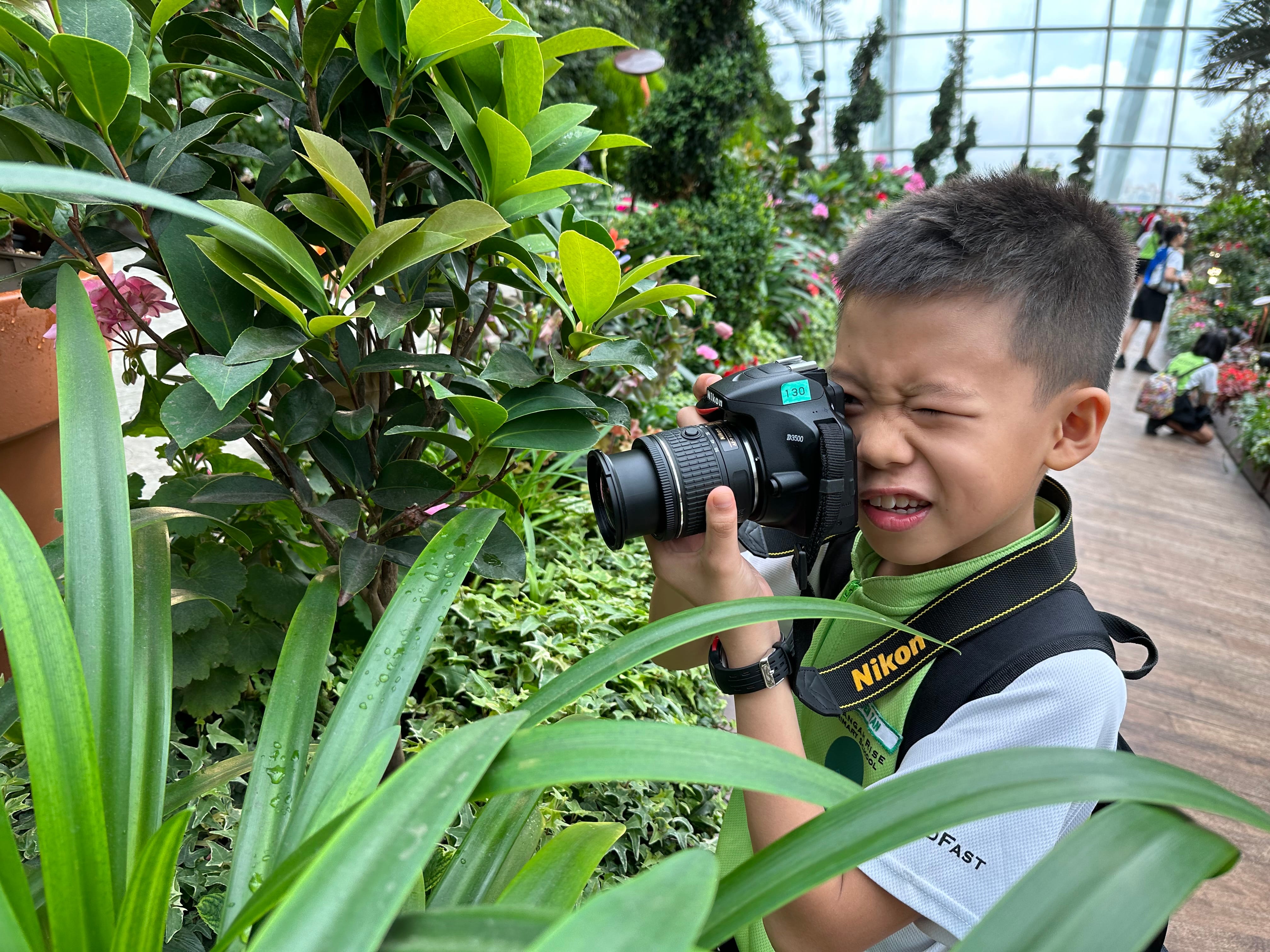 Boy squints while holding a Nikon camera near leafy plants in a greenhouse with flowers behind.